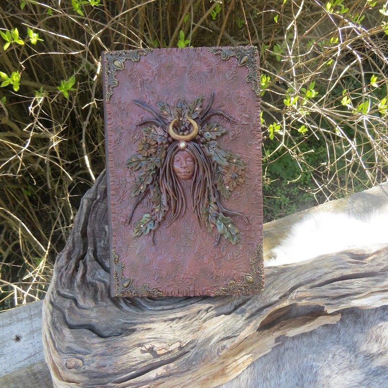 May include: A brown leather journal with a decorative embossed design. The cover features a sculpted image of a woman with a crown of leaves and a crescent moon.