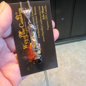 May include: A silver necklace with a glass vial pendant filled with dried herbs and a black gemstone. The pendant is attached to a silver chain and has a decorative silver cap. The necklace is displayed on a black card with the text "Way of the Cauldron" and contact information.