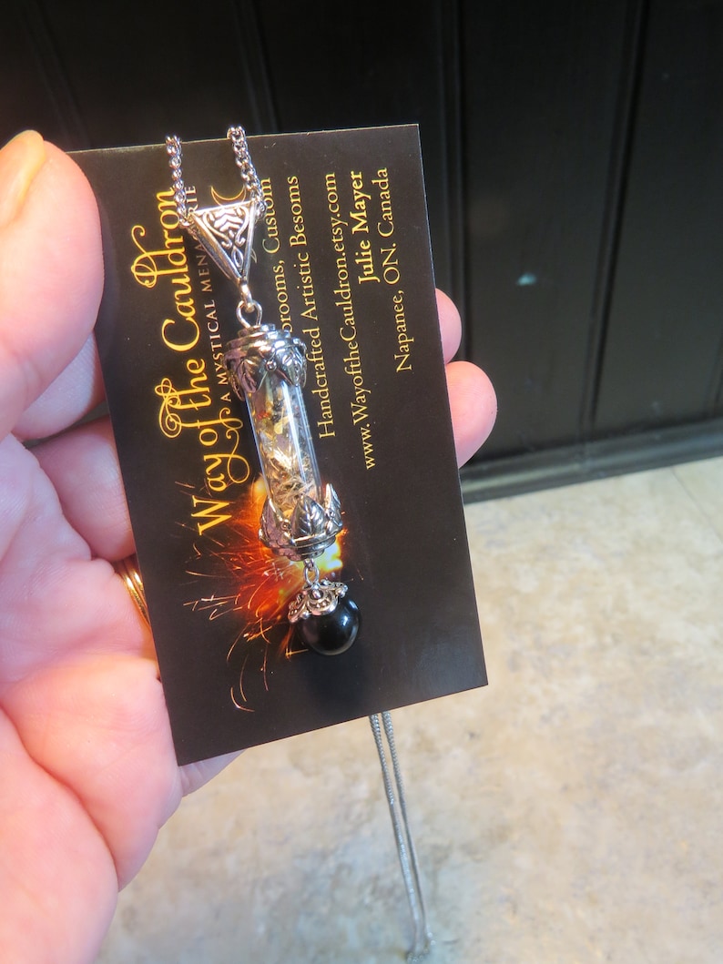 May include: A silver necklace with a glass vial pendant filled with dried herbs and a black gemstone. The pendant is attached to a silver chain and has a decorative silver cap. The necklace is displayed on a black card with the text "Way of the Cauldron" and contact information.