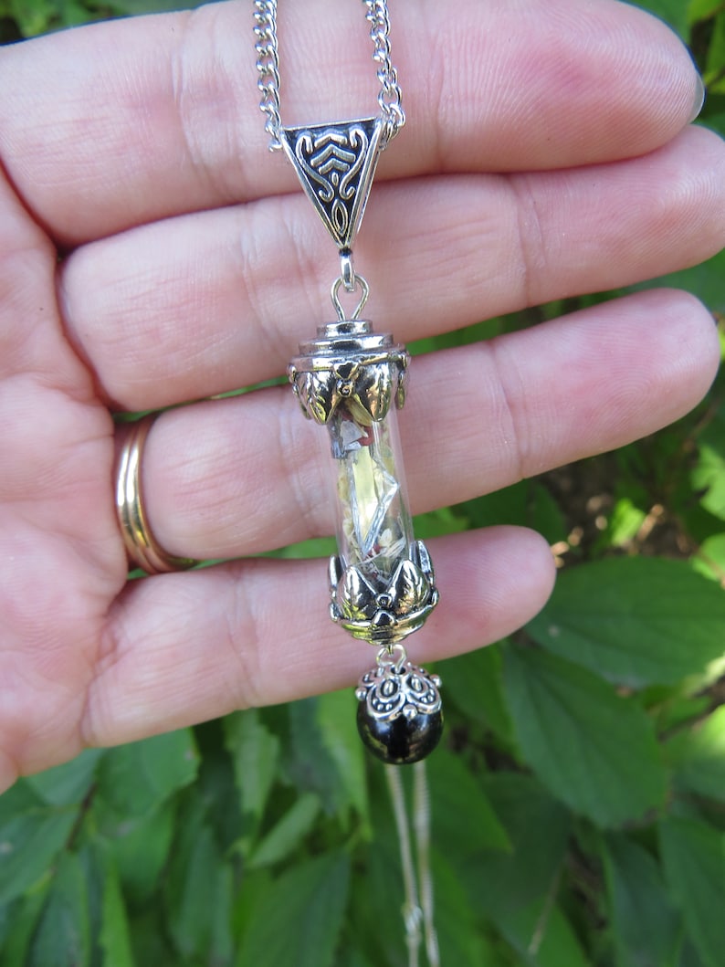 May include: A silver chain necklace with a pendant featuring a clear glass vial filled with small, sparkly objects. The vial is adorned with silver filigree and a black bead hangs from the bottom.