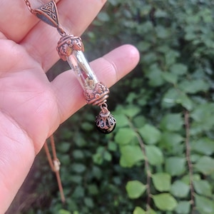 May include: A copper necklace with a glass vial pendant filled with dried botanicals and a black gemstone charm.
