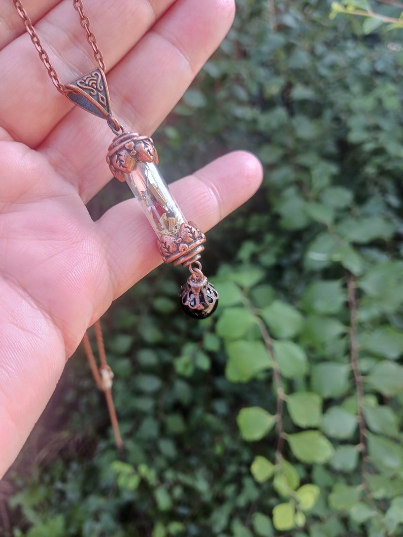 May include: A copper necklace with a glass vial pendant filled with dried botanicals and a black gemstone charm.