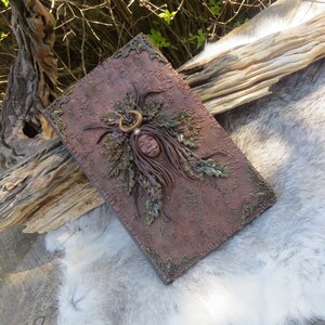 May include: A brown leather-bound journal with a decorative design featuring a woman's face surrounded by leaves and a crescent moon.