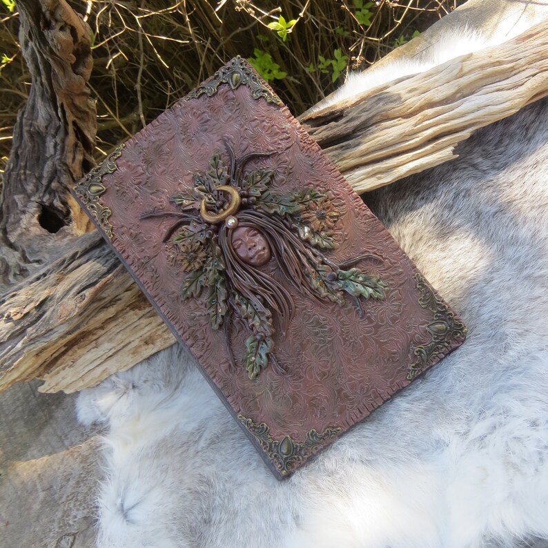 May include: A brown leather-bound journal with a decorative design featuring a woman's face surrounded by leaves and a crescent moon.