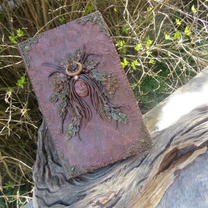 May include: A brown leather journal with a decorative design featuring a woman's face surrounded by leaves and flowers. The journal has gold accents and a crescent moon above the woman's head.