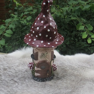 May include: A whimsical mushroom-shaped fairy house with a brown and white polka dot roof, a small door, and a dragonfly detail. The house is made of paper mache and is sitting on a white furry surface.