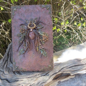 May include: A brown leather journal with a decorative embossed design. The cover features a sculpted image of a woman with a crown of leaves and a crescent moon.