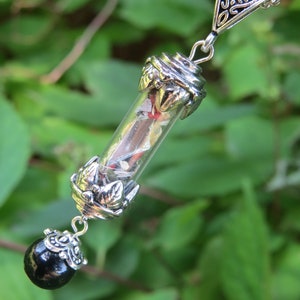 May include: A silver necklace with a glass vial pendant filled with colorful confetti and a black bead at the bottom. The pendant is attached to a silver chain with a triangular charm.