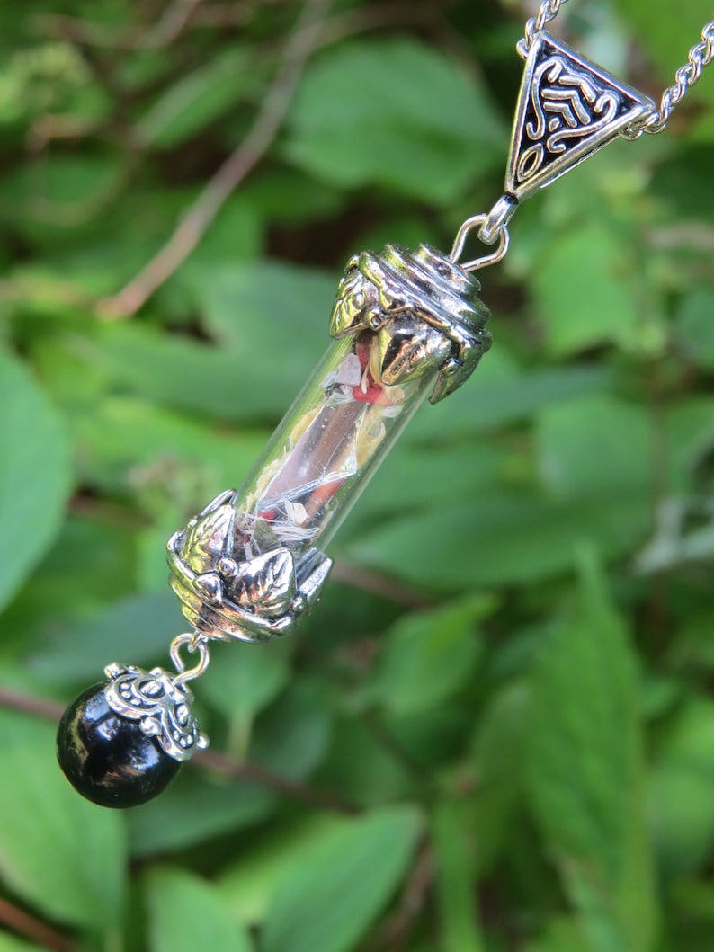 May include: A silver necklace with a glass vial pendant filled with colorful confetti and a black bead at the bottom. The pendant is attached to a silver chain with a triangular charm.