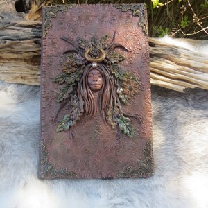 May include: A brown leather-bound journal with a decorative embossed design. The cover features a sculpted woman's face with a crown of leaves and a crescent moon.