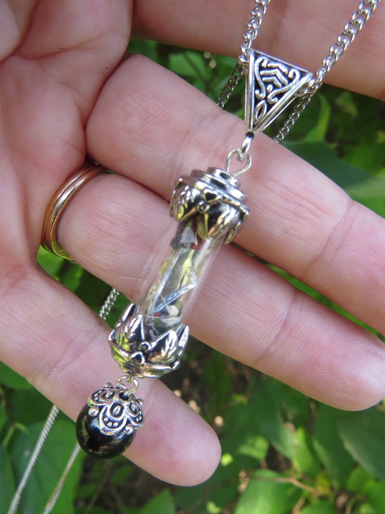 May include: A silver chain necklace with a glass vial pendant filled with dried flowers and herbs. The vial is attached to a silver charm with a black bead at the bottom.