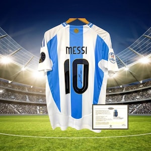 Puede incluir: Una camiseta de fútbol blanca y azul claro con el número 10 y el nombre "Messi" en la espalda. La camiseta está firmada por Messi y tiene un parche dorado en la manga.