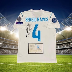 May include: White soccer jersey with the name "SERGIO RAMOS" in light blue, and the number "4" in blue. The jersey has a signature and a "Signed" certificate. The background shows a stadium.