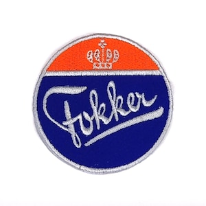 Puede incluir: Un parche circular azul y naranja con la palabra "Fokker" en letras cursivas blancas y una corona sobre la palabra.