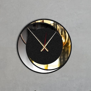 May include: A black and gold round wall clock with a mirrored face and black hands. The clock is hanging on a gray wall above a beige couch with pillows.