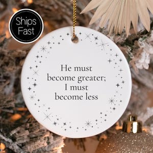 May include: A round white ceramic ornament featuring the black text 'He must become greater; I must become less' centered on the front. The design is framed by a delicate border of small black stars and sparkles, suspended by a gold string.