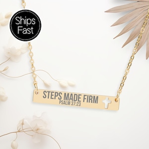 Op de afbeelding: Goudkleurige ketting met een rechthoekige hanger. De hanger heeft de woorden "STEPS MADE FIRM" en "PSALM 37:23" gegraveerd, samen met een kruisuitsparing. De ketting wordt gepresenteerd op een witte achtergrond met bloemaccenten.