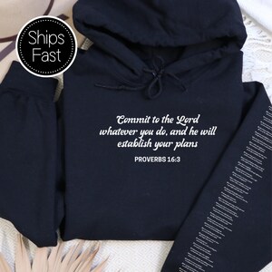 May include: Black hoodie featuring the white text "Commit to the Lord whatever you do, and he will establish your plans PROVERBS 16:3". The hoodie has a hood and long sleeves. The sleeve has white text. A white and black circle with the words "Ships Fast" is in the upper left corner.