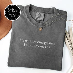 May include: A dark grey t-shirt with the quote "He must become greater; I must become less" printed in white. A silver necklace with a pendant lies on the shirt. The shirt is folded on a white surface.