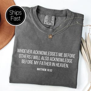May include: A charcoal gray Comfort Colors t-shirt with the text "WHOEVER ACKNOWLEDGES ME BEFORE OTHERS I WILL ALSO ACKNOWLEDGE BEFORE MY FATHER IN HEAVEN. MATTHEW 10:32" in white. A silver necklace with a pendant rests on the shirt.