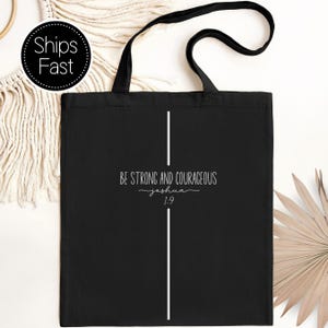 May include: Black tote bag with white text that reads "BE STRONG AND COURAGEOUS" and "joshua 1:9". The bag has a vertical white line design. The bag has black handles.
