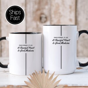 May include: Two white ceramic mugs with black handles and interiors. Each mug features a vertical black line and the black text: PROVERBS 17:22 A Cheerful Heart Is Good Medicine. The mugs are of different heights, displaying the same design.