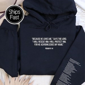 Op de afbeelding: Een zwarte hoodie met witte tekst uit Psalm 91:14. De tekst luidt: "OMDAT HIJ VAN MIJ HOUDT," ZEGT DE HEER, "IK ZAL HEM REDDEN; IK ZAL HEM BESCHERMEN, WANT HIJ ERKENT MIJN NAAM." De hoodie heeft een capuchon en lange mouwen.