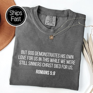 Könnte beinhalten: Ein anthrazitfarbenes T-Shirt mit Rundhalsausschnitt und weißem Text: But God demonstrates His own love for us in this while we were still sinners Christ died for us. Romans 5:8. Das T-Shirt hat ein Etikett der Marke Comfort Colors am Halsausschnitt.
