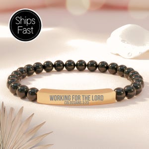May include: Black beaded bracelet with a gold-colored rectangular centerpiece. The centerpiece is engraved with the words "WORKING FOR THE LORD" and "COLOSSIANS 3:23". The bracelet is on a light pink surface.