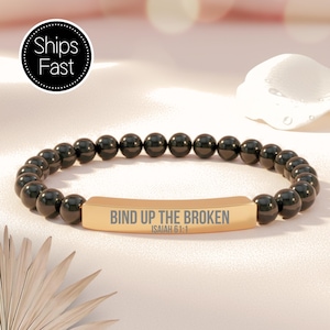 May include: Black beaded bracelet with a gold-colored rectangular centerpiece. The centerpiece is engraved with the words "BIND UP THE BROKEN" and the reference "ISAIAH 61:1". The bracelet is made of small, round, black beads.