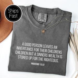 Könnte beinhalten: Ein anthrazitfarbenes Comfort Colors T-Shirt mit dem weißen Text "A GOOD PERSON LEAVES AN INHERITANCE FOR THEIR CHILDREN BUT A SINNERS WEALTH IS STORED UP FOR THE RIGHTEOUS. PROVERBS 13:22". Eine silberne Halskette mit Anhänger liegt auf dem Shirt.