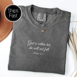 Könnte beinhalten: Ein graues Comfort Colors T-Shirt mit dem Text "God is within her, she will not fall" und Psalm 46:5 in Weiß gedruckt. Eine silberne Halskette mit Anhänger liegt auf dem Shirt. Das Shirt ist gefaltet.