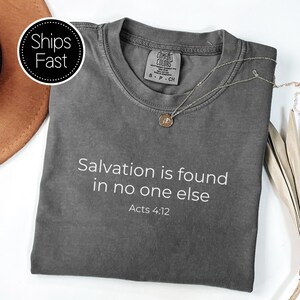 Peut inclure: Un t-shirt gris avec le texte "Salvation is found in no one else" et "Acts 4:12" imprimé en blanc. Un collier en argent avec un pendentif circulaire repose sur le t-shirt. Le t-shirt est plié sur une surface blanche.