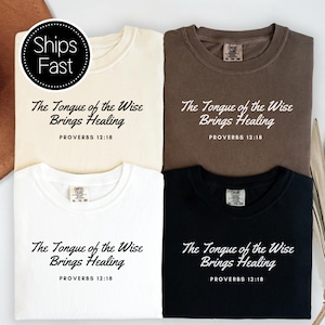 May include: Four crew neck t-shirts in cream, brown, white, and black. Each shirt features the text "The Tongue of the Wise Brings Healing" in a script font, followed by "PROVERBS 12:18" in a sans-serif font. The shirts have a visible brand label.