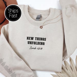 May include: A cream-coloured sweatshirt featuring the text "NEW THINGS UNFOLDING Isaiah 43:19" in black. A gold necklace with a small pendant is visible. The sweatshirt is folded, with a portion of a hat in the background.