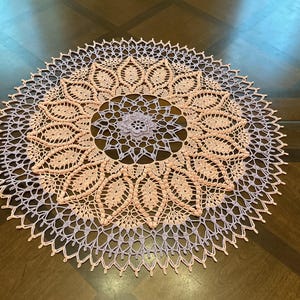 May include: A round, hand-crocheted doily featuring a central lavender flower motif surrounded by concentric rings of peach-colored leaf patterns and an outer border of delicate lavender lace stitching in a repeating geometric design.