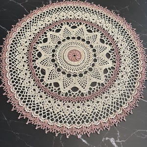 Puede incluir: Un tapete redondo de ganchillo color crema con un diseño calado delicado. El tapete presenta un borde rosa y un motivo floral rosa central. Este artículo hecho a mano es perfecto para añadir un toque de encanto vintage a cualquier decoración del hogar.