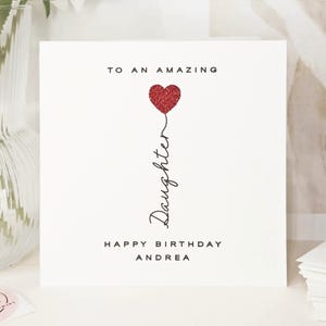 Personalised name daughter birthday card with the message ‘TO AN AMAZING Daughter HAPPY BIRTHDAY Andrea’. The word ‘Daughter’ is written vertically in cursive, with a red glitter heart. Styled on a white surface with a beige vase of white flowers and a stack of books in the background, creating a soft and elegant scene.