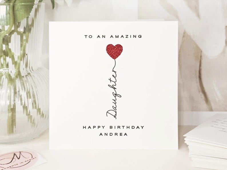 Personalised name daughter birthday card with the message ‘TO AN AMAZING Daughter HAPPY BIRTHDAY Andrea’. The word ‘Daughter’ is written vertically in cursive, with a red glitter heart. Styled on a white surface with a beige vase of white flowers and a stack of books in the background, creating a soft and elegant scene.
