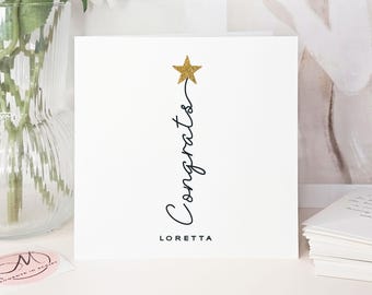 May include: A white greeting card featuring the word "Congrats" in black script, with a gold glitter star. The name "Loretta" is printed below. A stack of white cards is visible in the background.