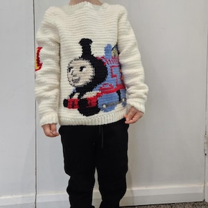 May include: A white crocheted jumper with a Thomas the Tank Engine design. The jumper has long sleeves and a crew neck.