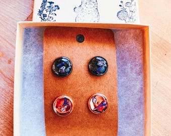 May include: Four stud earrings presented in a gift box. Two earrings feature a dark background with blue and green details, and two have a red and orange bird design. The box is labelled "BLUE BONNET FARM" with a dog illustration.