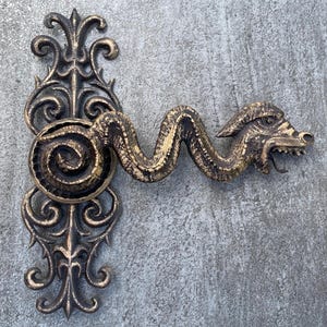 Puede incluir: Una manija de puerta decorativa con un diseño de dragón. La manija está hecha de metal y tiene un acabado marrón oscuro. La cabeza del dragón está orientada hacia la derecha y tiene un diseño detallado. La manija está unida a un trabajo de volutas decorativo.