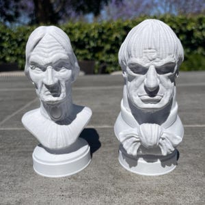 May include: Two white busts, one depicting an elderly person with a stern expression and the other a man with a bow tie. Both are on round bases and appear to be 3D-printed. The busts are set against a blurred outdoor background.