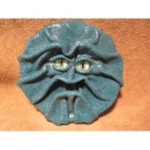 May include: A teal-colored, round, sculpted face with a textured surface and a grumpy expression. The face has two green, reptilian-like eyes and a furrowed brow. The mouth is slightly open, and the overall design is reminiscent of a monster or creature.