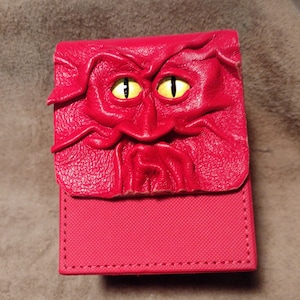 May include: A rectangular red deck box featuring a sculpted leather-like flap with a textured, wrinkled face design. The face has two prominent yellow eyes with black vertical pupils. The base of the box is made of a solid, textured red material.