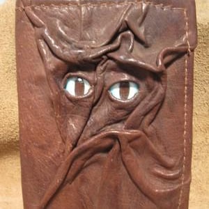 May include: Brown leather wallet with a sculpted face design. The face has two white and brown eyes and is made to look like a monster. The wallet is rectangular and has stitched edges.