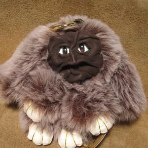 May include: A plush keychain featuring a dark brown, wrinkled, sculpted face with visible eyes surrounded by thick, mauve-colored faux fur. The base of the keychain includes four white, padded, paw-like appendages with gold-colored stitching details.
