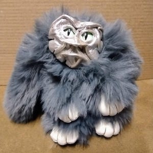 May include: A plush creature featuring long, dark gray faux fur, a metallic silver face with sculpted features, and green eyes. The figure has white, padded paws extending from the fur, creating a textured appearance with distinct facial details.