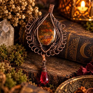 May include: A copper wire-wrapped pendant featuring a central oval-shaped moss agate stone with earthy green, brown, and orange inclusions. A faceted, teardrop-shaped red crystal bead hangs from the bottom of the intricate metal frame.